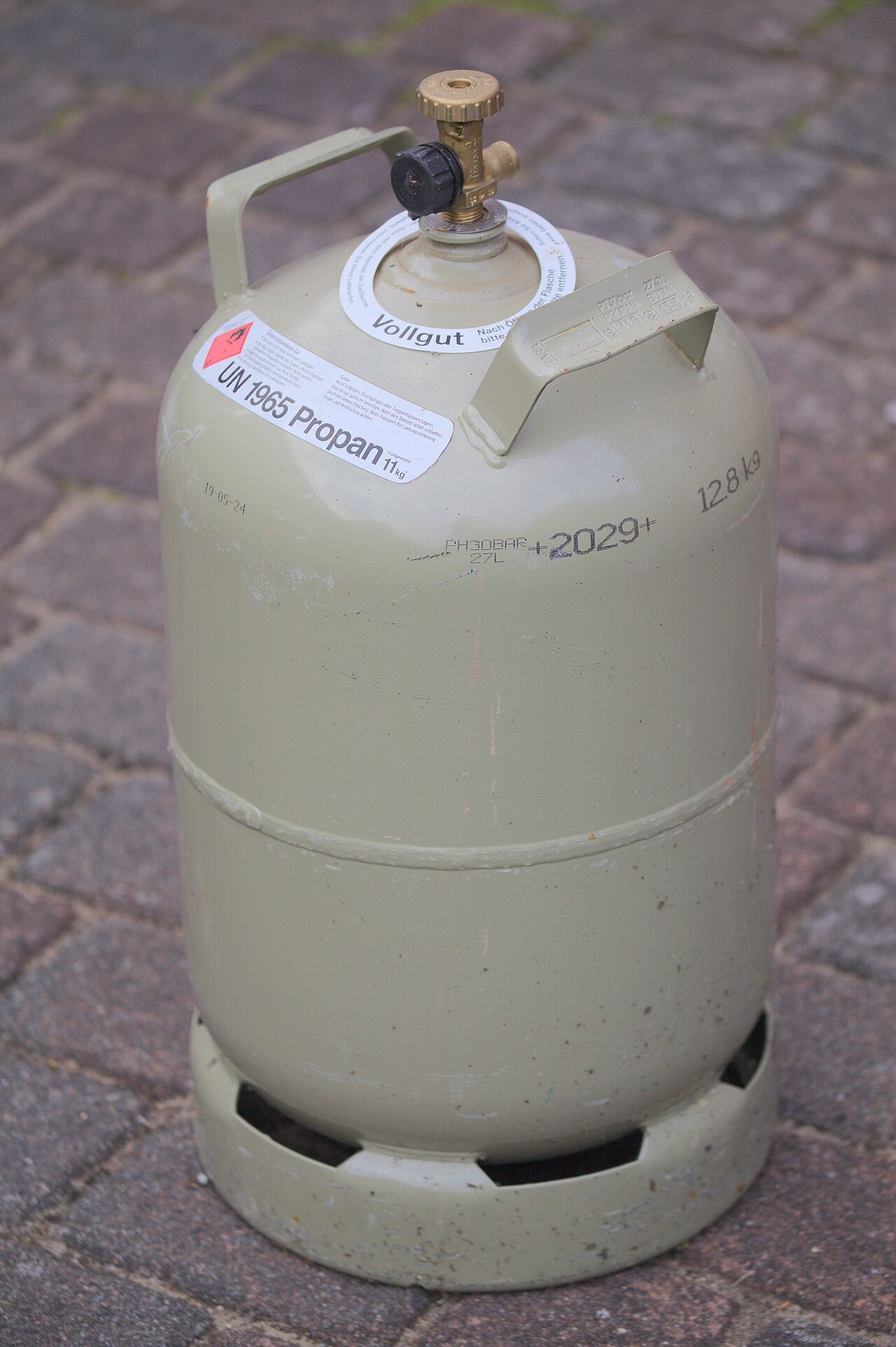 A grey 11 kg propane gas cylinder with a valve and a safety label, standing on a brick pavement