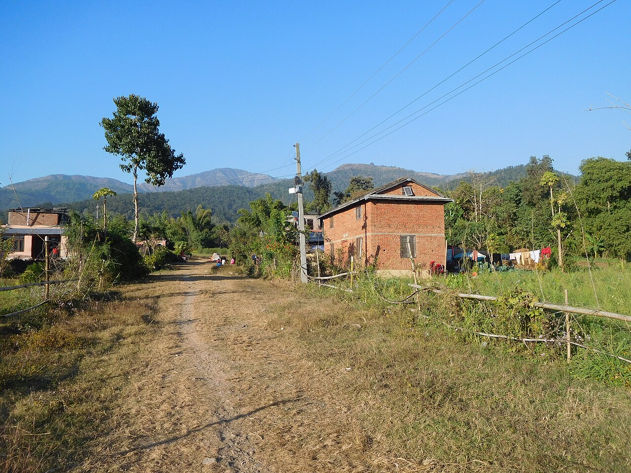 A two-storey red-brick house beside a dirt path, with green hills behind, in Surkhet, Nepal